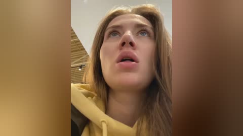 Media: A video of a young woman with light skin, blue eyes, and straight brown hair, wearing a mustard-yellow hoodie, looking up with an expression of surprise or fear. The background is blurry.