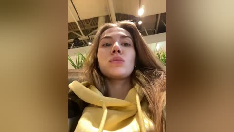 Media: Video of a young woman with long, light brown hair wearing a yellow hoodie, taken indoors with a modern, industrial ceiling and green plants in the background.