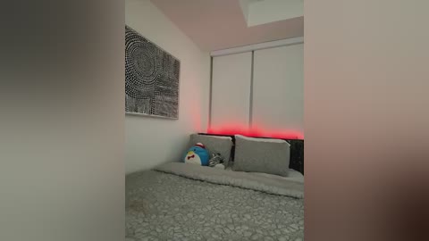 Media: Video of a minimalist bedroom with a plush grey carpet, beige walls, a black and white mandala tapestry, and a bed with grey pillows and a plush toy. A red light under a closed window adds a soft glow.