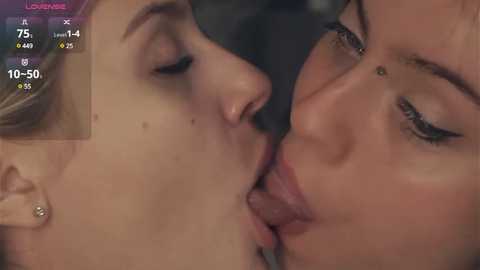 Media: Video: Two women, one with blonde hair, the other with brown hair, are passionately kissing. Their faces are close, with the blonde woman's tongue extended. Background is blurred.