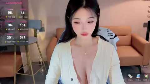 Media: A video of an East Asian woman with long black hair, fair skin, and large breasts, wearing a low-cut white cardigan, seated in a modern, cozy living room.