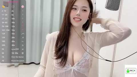 Media: Video of an East Asian woman with long brown hair, wearing a sheer white blouse revealing a lacy bra, smiling while adjusting her hair. Background includes a calendar, gray curtains, and a speaker.