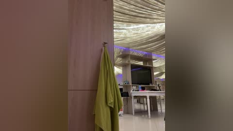 Media: Video of a modern living room with beige walls, a green towel hanging on a wooden door, a white dining table, and a ceiling with draped fabric.