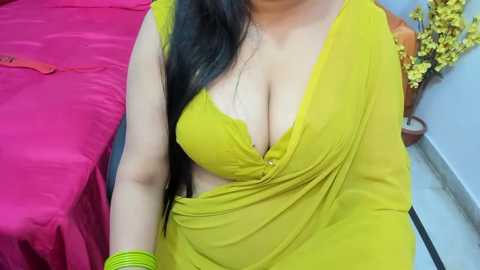 Media: Video of a South Asian woman with light skin, long black hair, wearing a yellow saree with a deep V-neck, revealing ample cleavage, seated on a bed with red sheets.