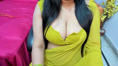 Media: Video of a South Asian woman with medium skin tone, wearing a bright yellow saree with a low-cut blouse revealing ample cleavage. She has long, straight black hair. Background features a red bedspread, a potted plant, and a tiled floor.