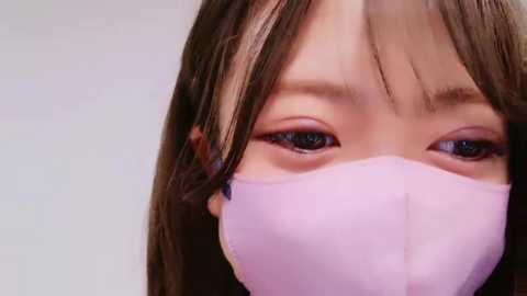 Media: Close-up video of an Asian woman with long, straight brown hair, wearing a pink face mask that covers her nose and mouth. Her large, expressive brown eyes are partially visible. The background is a plain, light-colored wall, emphasizing her face.