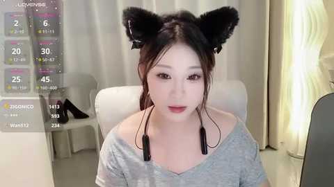 Media: Video of an East Asian woman with light skin, brown hair, and black cat ears, wearing a grey off-shoulder top, seated indoors with white curtains and a digital display showing weather information.