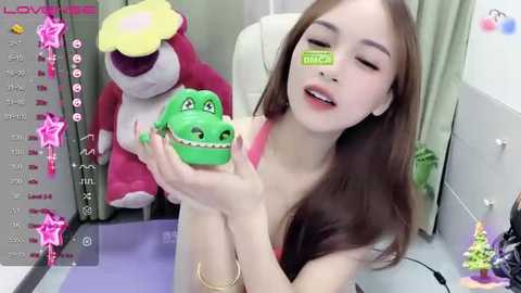 Media: A young, fair-skinned woman with long brown hair, wearing a low-cut pink top, holds a green frog cake with a green band-aid on her nose, surrounded by plush toys in a colorful room.