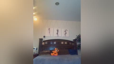 Media: Video of a dimly lit bedroom featuring a dark wooden bed with a plush orange bear on a blue bedspread, framed black-and-white dancer silhouette artwork on the wall, and a small white lamp on the left.