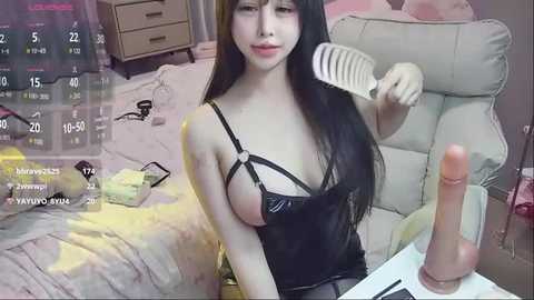 Media: A video of a young Asian woman with long black hair, wearing black lingerie, sitting in a chair in a bedroom. She holds a hairbrush and a large, erect penis in her other hand.