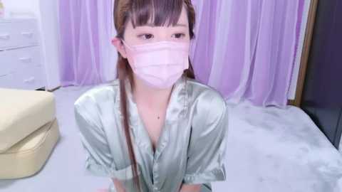 Media: A video of a young Asian woman with light skin, wearing a light pink surgical mask, seated on a white floor. She has long brown hair, straight bangs, and is dressed in a shiny, light green satin robe. The background features light purple curtains and a white dresser with drawers.