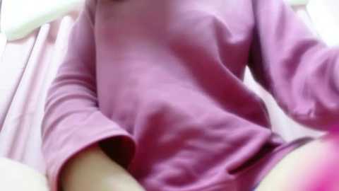 Media: A video of a person wearing a loose, long-sleeved pink shirt with a relaxed fit, sitting with legs apart, revealing light-colored underwear. The background is out of focus, possibly indoor, with soft, diffused lighting.