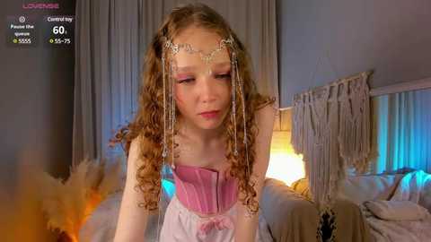 Media: Video of a young girl with curly brown hair, wearing a pink corset and silver headpiece, standing in a cozy, dimly lit bedroom with beige curtains and a warm, yellow-lit lamp.