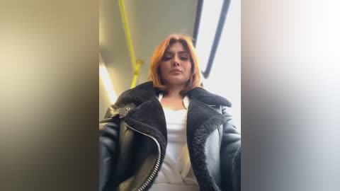 Media: A video of a young woman with light skin and shoulder-length, straight orange hair, wearing a black leather jacket and a white top, standing in a subway car. The background is dimly lit with yellow lighting.