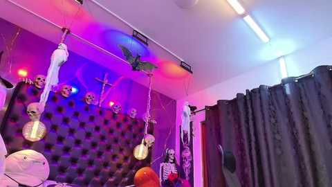 Media: Video of a brightly lit, Halloween-themed room with a black, tufted headboard, skeletons, and a bat hanging from the ceiling. Purple and pink lights add ambiance, while a black curtain hangs in the background.