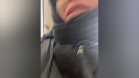 Media: A close-up video of a person's lower face and neck, wearing a black puffer jacket with a hood, partially covering their mouth. The background is blurred, suggesting an indoor setting.