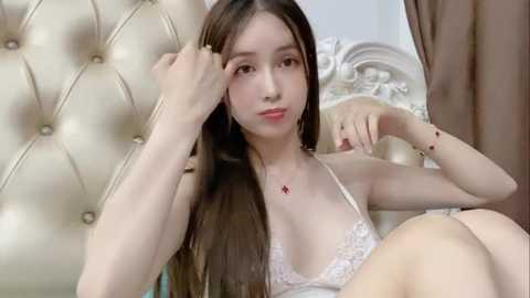 Media: Video of a young Asian woman with long brown hair, fair skin, and small breasts, wearing a white lace bra, sitting on a cream-colored tufted leather sofa, touching her hair.