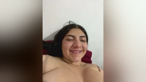 Media: A video of a smiling, topless woman with medium breasts and medium skin tone, lying on a red pillow against a white wall. Her dark hair is tied up, and she has a gold necklace.