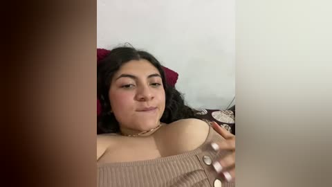 Media: A video of a plus-size woman with curly black hair, wearing a beige off-shoulder top, lying on a bed with a red pillow and patterned blanket, smiling while holding her breast.