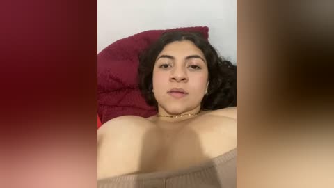 Media: Video of a young woman with medium skin tone, dark curly hair, and light makeup, lying on a bed with red and brown pillows. She's wearing a beige top.