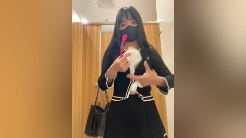 Media: Video of an East Asian woman in a black schoolgirl outfit with a face mask, holding a pink spoon, standing in a beige hallway with wooden doors.