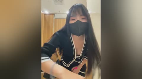 Media: Video of an Asian woman with long black hair, wearing a black mask, black top, and white gloves, standing in a modern, beige-walled room with a brown curtain and a ceiling light fixture.