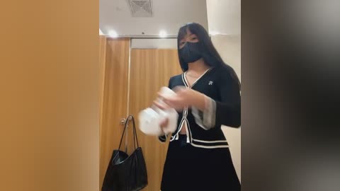 Media: Video of an Asian woman wearing a black face mask and navy sailor uniform with white trim, holding a white plastic bottle, standing in a beige-tiled hallway with a brown curtain and a black handbag.