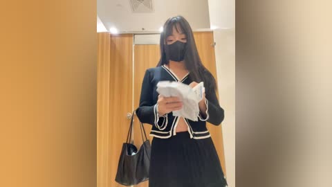 Media: Video of a young Asian woman in a black sailor-style outfit, black mask, reading a paper in a brightly lit, beige-walled hallway with a wooden door.