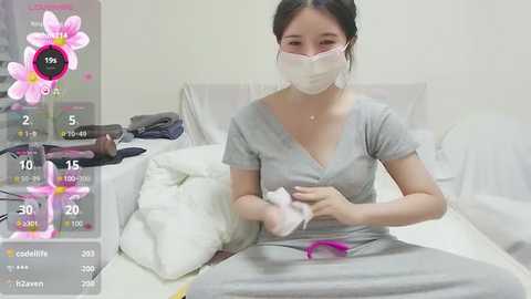 Media: Video of a young Asian woman in a hospital bed, wearing a mask, gray shirt, and pants, holding a white cloth. Background includes white sheets, a monitor, and a pink flower overlay.