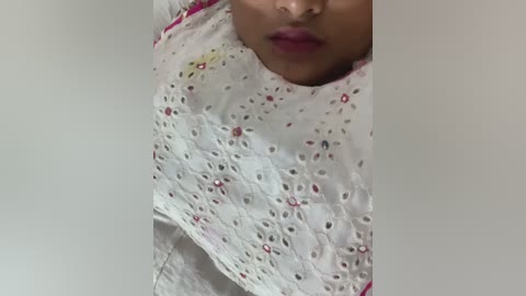 Media: Video of a young South Asian woman with medium brown skin, wearing a white lace dress with floral embroidery, standing in front of a plain white wall.