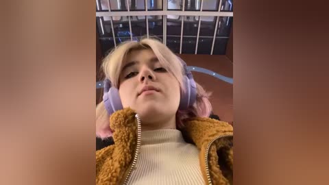 Media: Video of a young woman with light skin, blonde hair, and pink highlights, wearing a fuzzy brown jacket over a cream-colored sweater, lying on a metal bed in a hospital setting.