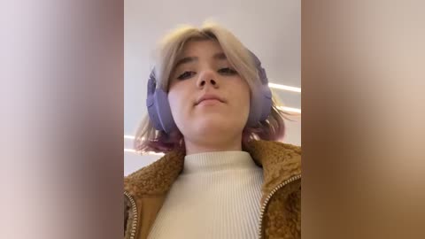 Media: Video of a young woman with blonde hair, wearing purple headphones, a beige fur jacket, and a white turtleneck sweater. The background is blurred, with a soft focus on her face, creating a candid, intimate atmosphere.