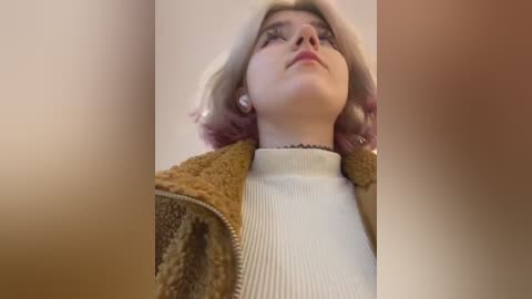 Media: Video of a young woman with light skin and shoulder-length, wavy pink hair. She wears a cream sweater and a brown, textured jacket. The background is blurred, with soft, warm tones.