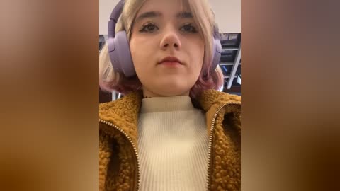 Media: A video of a young woman with light skin, blonde hair, and large purple headphones, wearing a brown textured jacket over a white turtleneck.