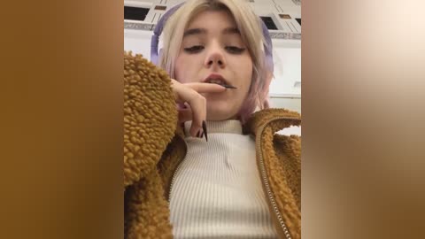 Media: Video of a young woman with platinum blonde hair, wearing a fuzzy brown jacket over a white ribbed sweater. She is biting her finger, with a neutral expression. Background shows a tiled ceiling and fluorescent lights.