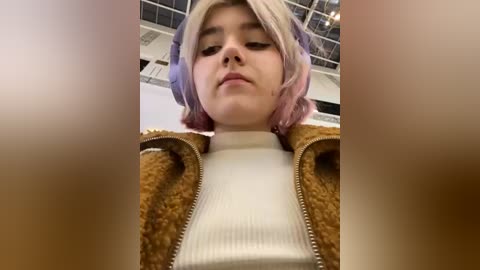 Media: Video of a young woman with short, dyed pink and purple hair, wearing a beige fur-lined jacket over a cream ribbed sweater, indoors with a metallic ceiling and air conditioning vents in the background.