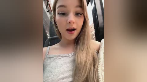 Media: Video of a young Caucasian girl with light skin, long blonde hair, and a surprised expression, wearing a light gray tank top, in a car.