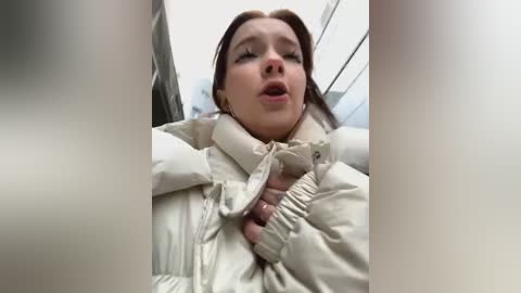 Media: A video of a woman in a white puffer jacket, arms pinned behind her back, looking distressed. The background shows a blurred, indoor setting with metallic railings.