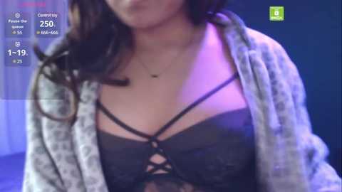 Media: A video of a woman with medium skin tone and dark hair, wearing a black lace bra with crisscross straps, over a light grey cardigan, indoors with purple lighting.