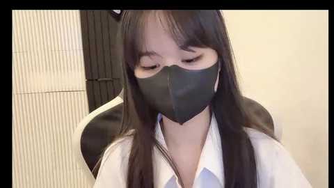 Media: Video of an East Asian woman with long black hair, wearing a black face mask, white blouse, and a black and white chair, indoors.