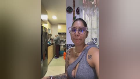 Media: A video of a woman with glasses, tattoos, and a grey tank top, standing in a modern kitchen with wooden cabinets and a sink.