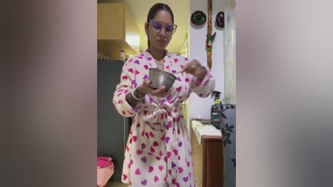 Media: A video of a middle-aged woman with glasses, wearing a pink heart-patterned robe, holding a bowl, in a cozy kitchen with wooden cabinets and holiday decorations.