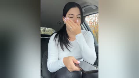 Media: Video of a young woman with long black hair, wearing a white long-sleeve top, sitting in a car, covering her face with her hand, looking upset.