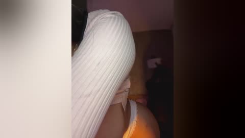 Media: Video of a woman with long, straight white hair, partially covering her face, wearing a white sleeveless top. Background shows a dimly lit room with a blurred figure.