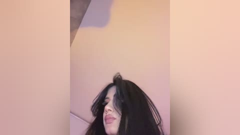 Media: Video of a woman with long black hair, pale skin, and slightly parted lips, standing indoors with a beige wall and a ceiling fan in the background.