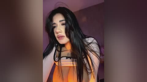 Media: Video of a young woman with long, straight black hair, wearing a white top, and looking down with a thoughtful expression. The background is dimly lit with purple and orange hues.