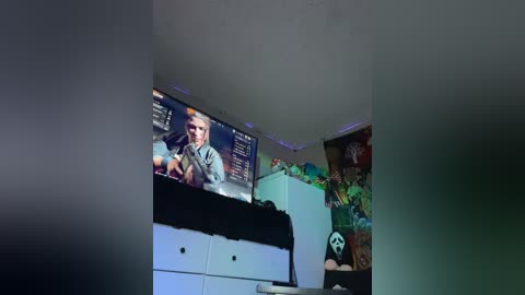 Media: Video of a dimly lit, cluttered bedroom with a TV displaying a video game, a white dresser, a plush panda toy, and a colorful wall mural.