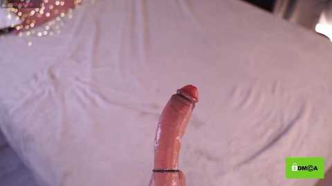 Media: Video of a large, erect penis on a white bedspread. The penis is circumcised, with visible veins and a slight glisten. The background is blurred, showing a hint of a pink, glittery object.