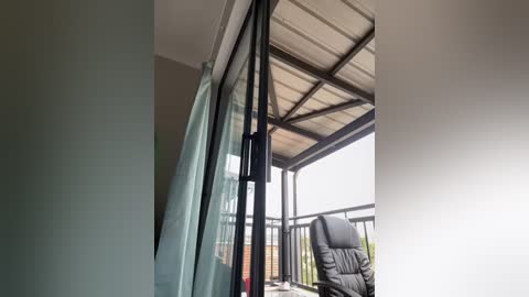 Media: Video of a modern balcony with glass railings, a black cushioned lounge chair, and a light blue curtain partially drawn, featuring a wooden pergola and a brick wall in the background.