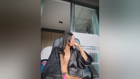 Media: Video of a Black woman with long, wavy hair, wearing a black satin robe, seated in a black chair. Background features modern, minimalist interior design with large windows and abstract art.